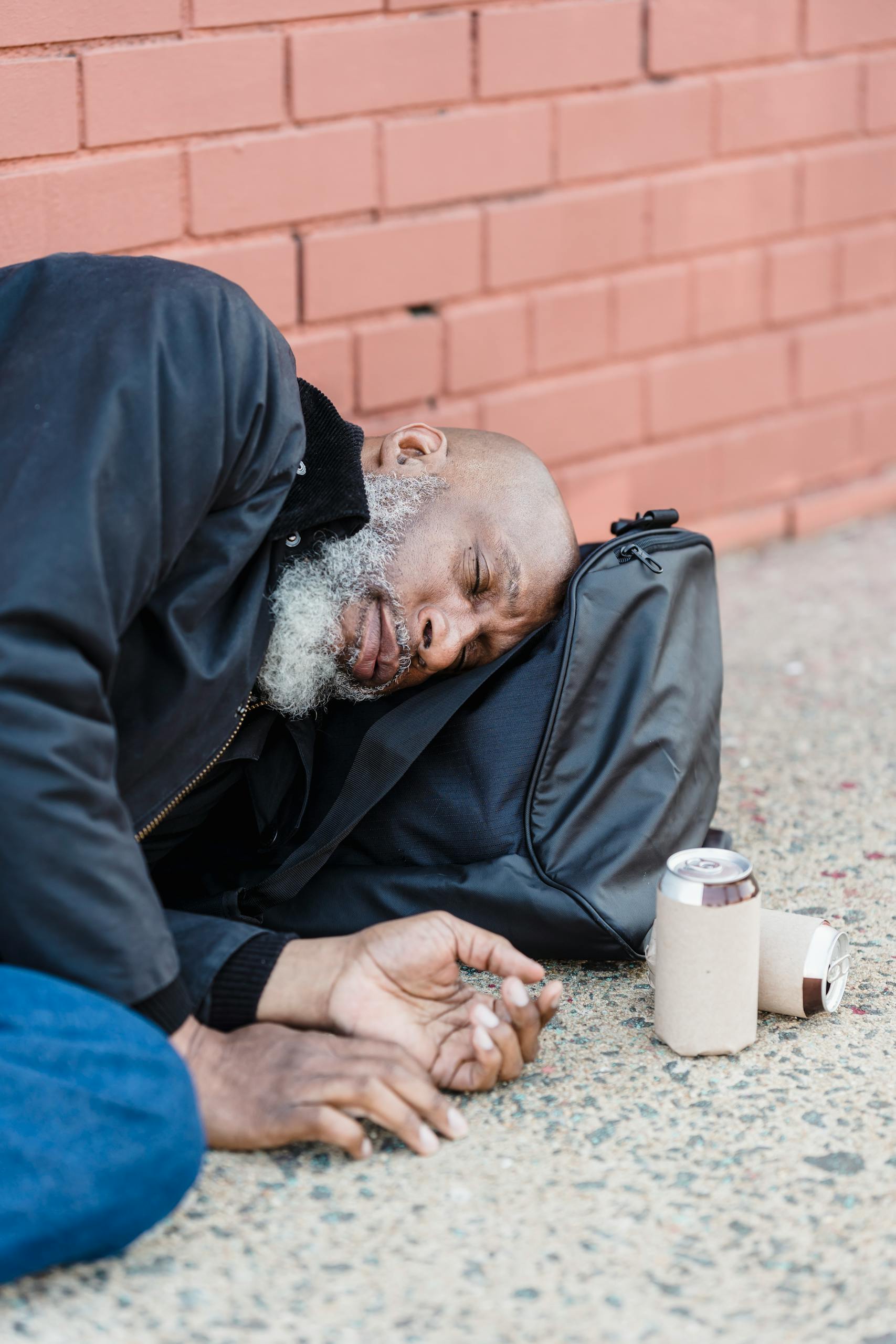 An elderly man rests on a sidewalk, leaning against a brick wall with a can beside him.