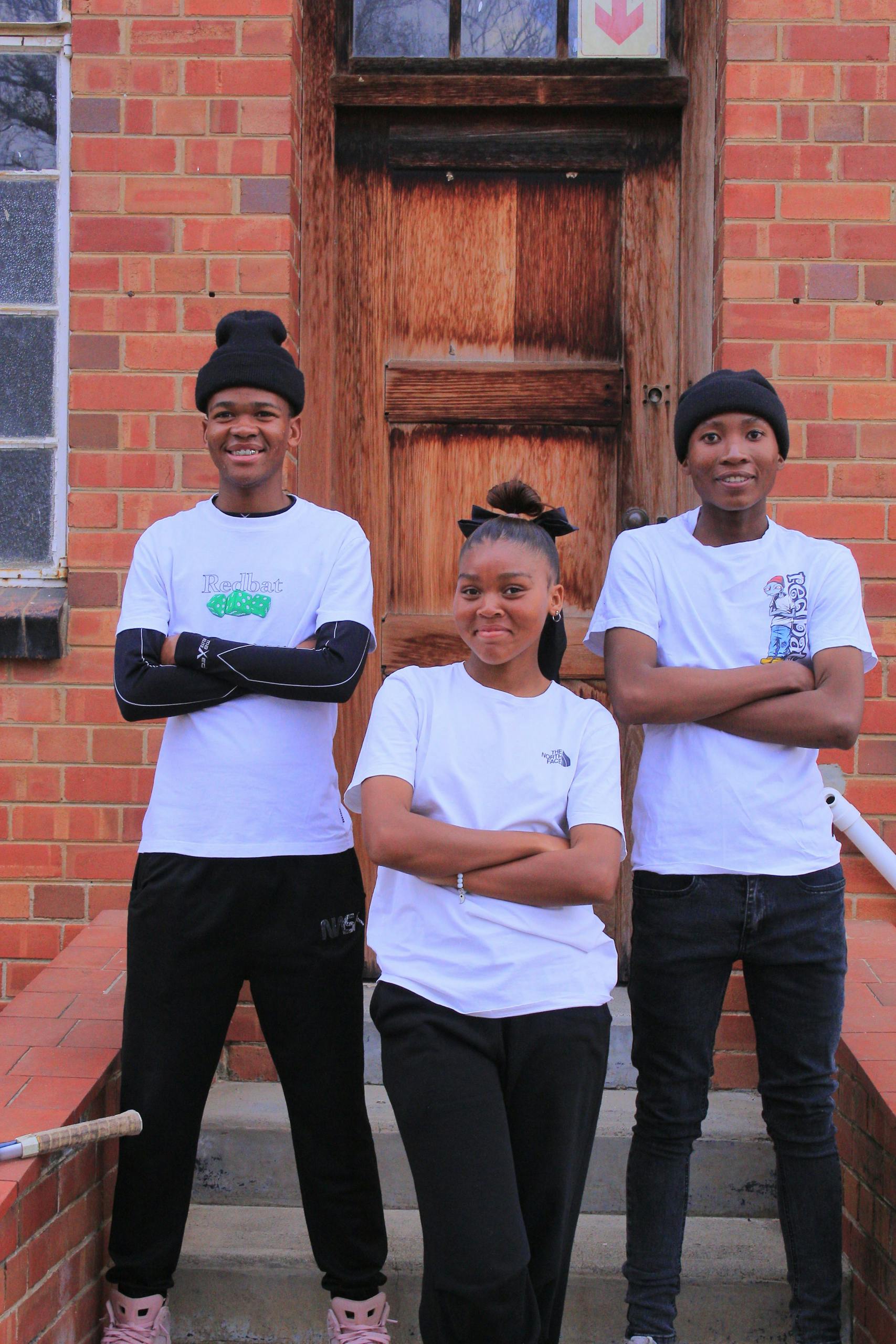 Three teenagers pose confidently in casual attire against a brick wall.